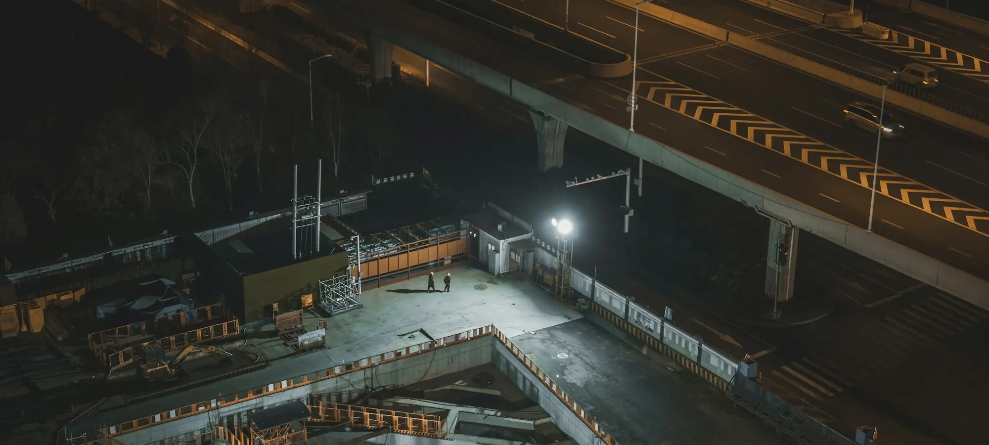 A construction site under floodlights at night beside an elevated highway